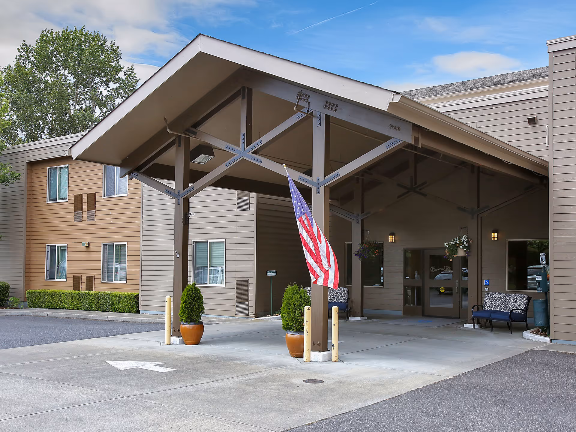 Entrance of a senior living facility with a covered drop-off area supported by brown beams. Two potted plants and an American flag are placed near the entrance. The building exterior is beige with multiple windows, and there are benches near the glass doors under the covered area.