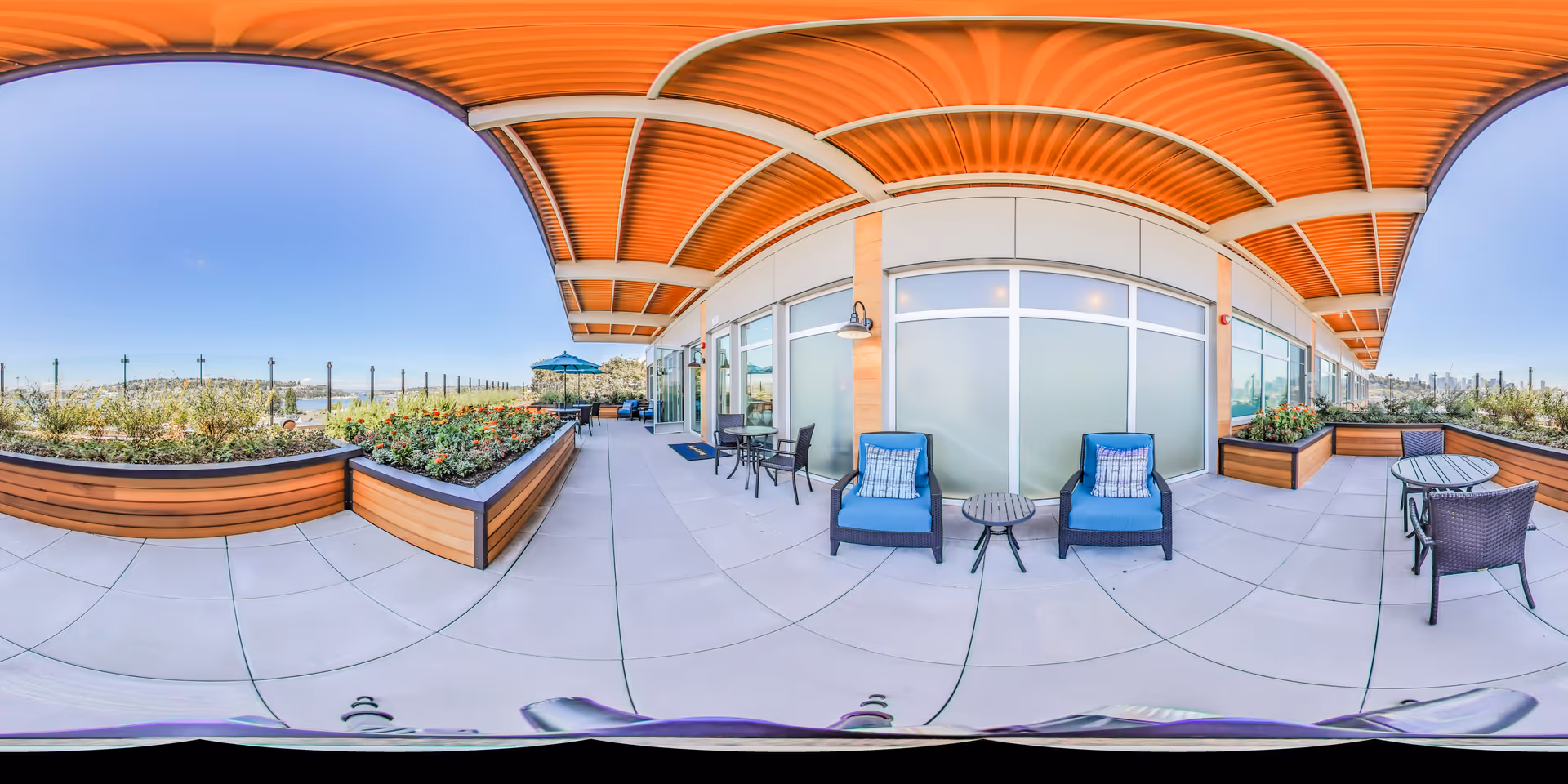 Outdoor patio area at Aegis Living Lake Union featuring a covered walkway with wooden ceiling, several seating arrangements including blue cushioned chairs and small tables, planter boxes with flowers, and a clear blue sky in the background.
