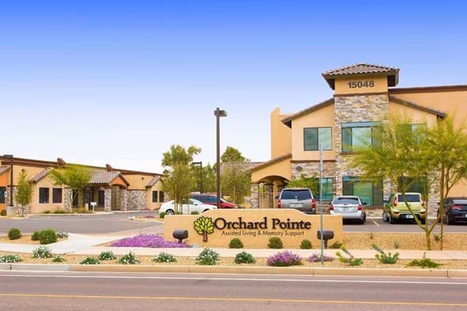 Exterior view of Orchard Pointe Assisted Living & Memory Support facility with a stone and stucco building, several parked cars, desert landscaping with small trees and flowering plants, and a clear blue sky.