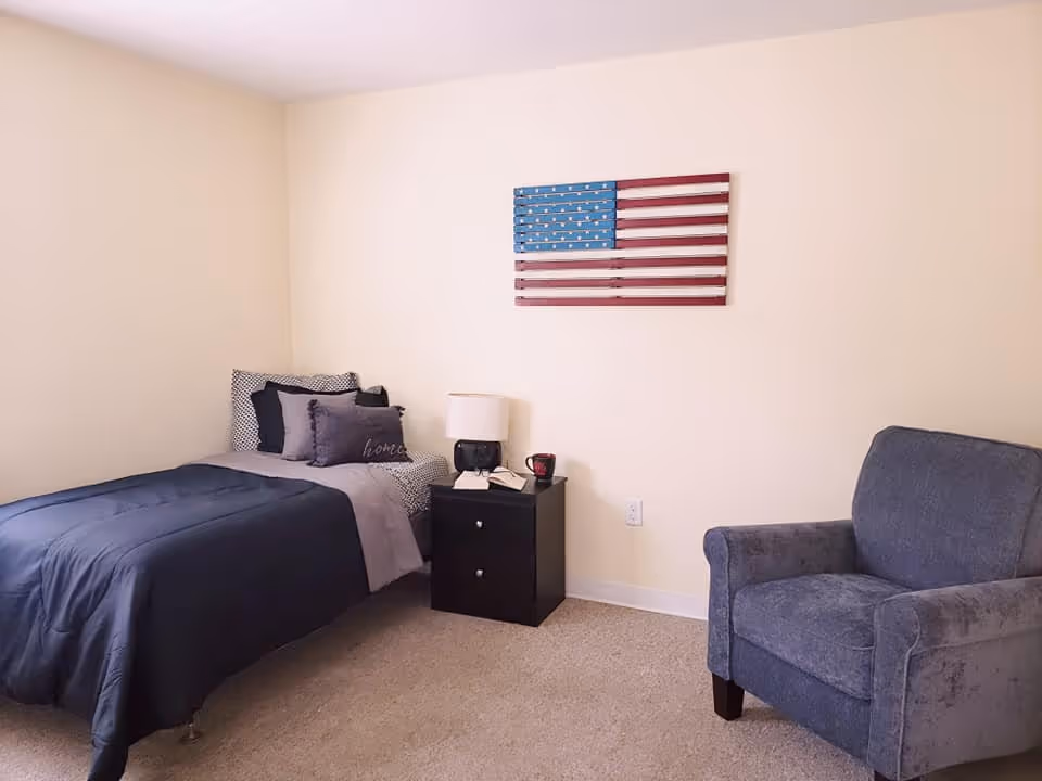 A simple bedroom with a single bed, bedside table and lamp, an armchair, and an American flag wall decoration.