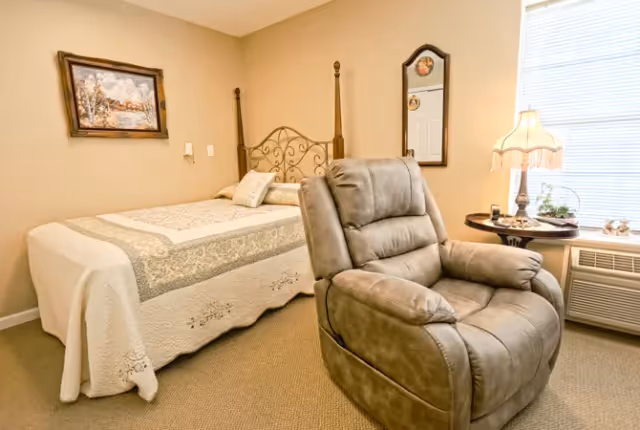 A cozy bedroom featuring a bed with a decorative quilt and pillows, a comfortable gray recliner chair in the foreground, a small round side table with a vintage-style lamp, and a window with blinds letting in natural light. A framed painting and a mirror hang on the beige walls.