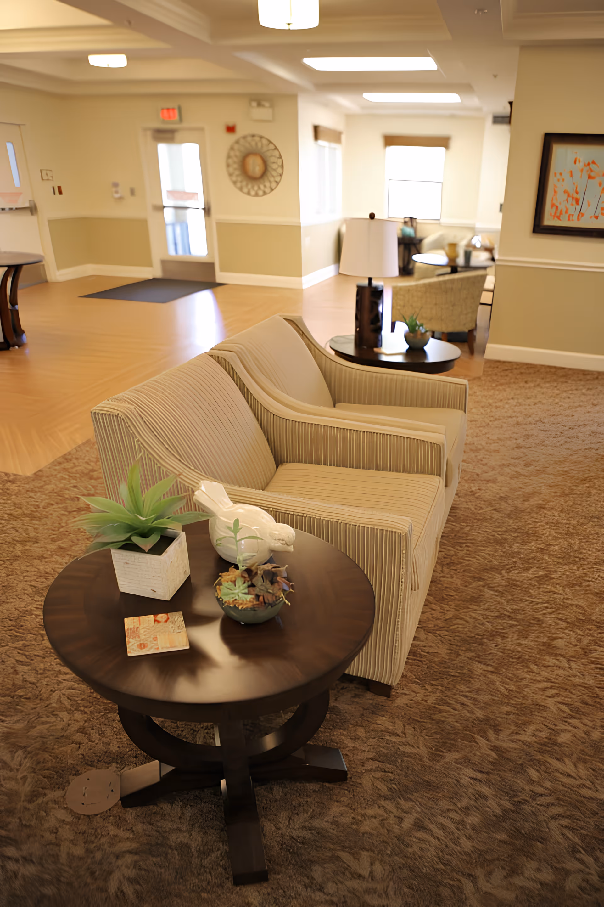 A cozy seating area in a senior living facility with a beige striped loveseat and a round wooden side table. The table holds a small potted plant, a decorative white bird, and a small bowl with dried leaves. In the background, there is another seating area with chairs, a table, a lamp, and a framed artwork on the wall. The room has beige walls, carpeted flooring, and large windows letting in natural light.