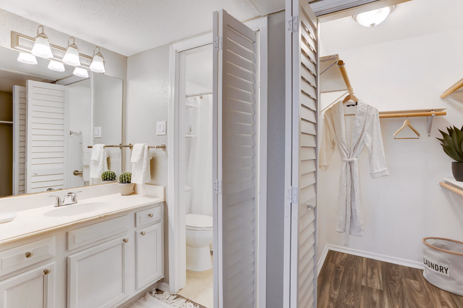 A bright bathroom with a white vanity, sink, and large mirror with three light fixtures above. To the right, there is a small closet area with wooden shelves, a hanging white bathrobe, a laundry basket, and a potted plant. The bathroom also has a separate toilet and shower area visible through an open door.