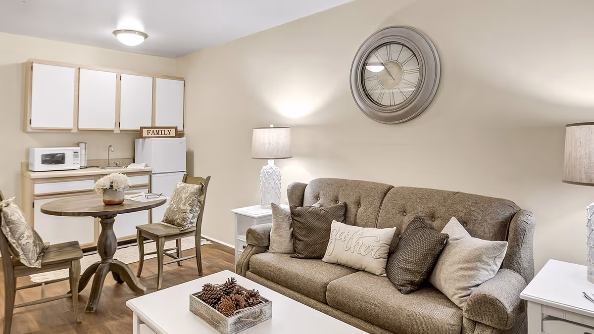 A cozy living area with a brown sofa adorned with multiple pillows, a white coffee table with a decorative tray of pinecones, and a round wooden dining table with two chairs. The background features a small kitchenette with white cabinets, a microwave, a sink, and a refrigerator. A large wall clock and two table lamps provide warm lighting.