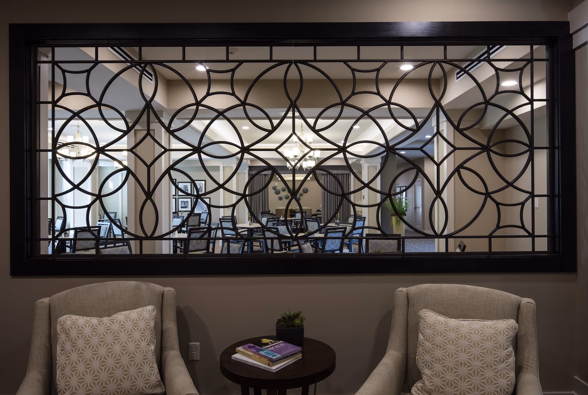 View through a decorative black metal screen into a dining area with multiple tables and chairs. In the foreground, two beige armchairs with patterned cushions flank a small round table holding books and a small plant. The dining area is well-lit with chandeliers and has neutral-colored walls and carpet.