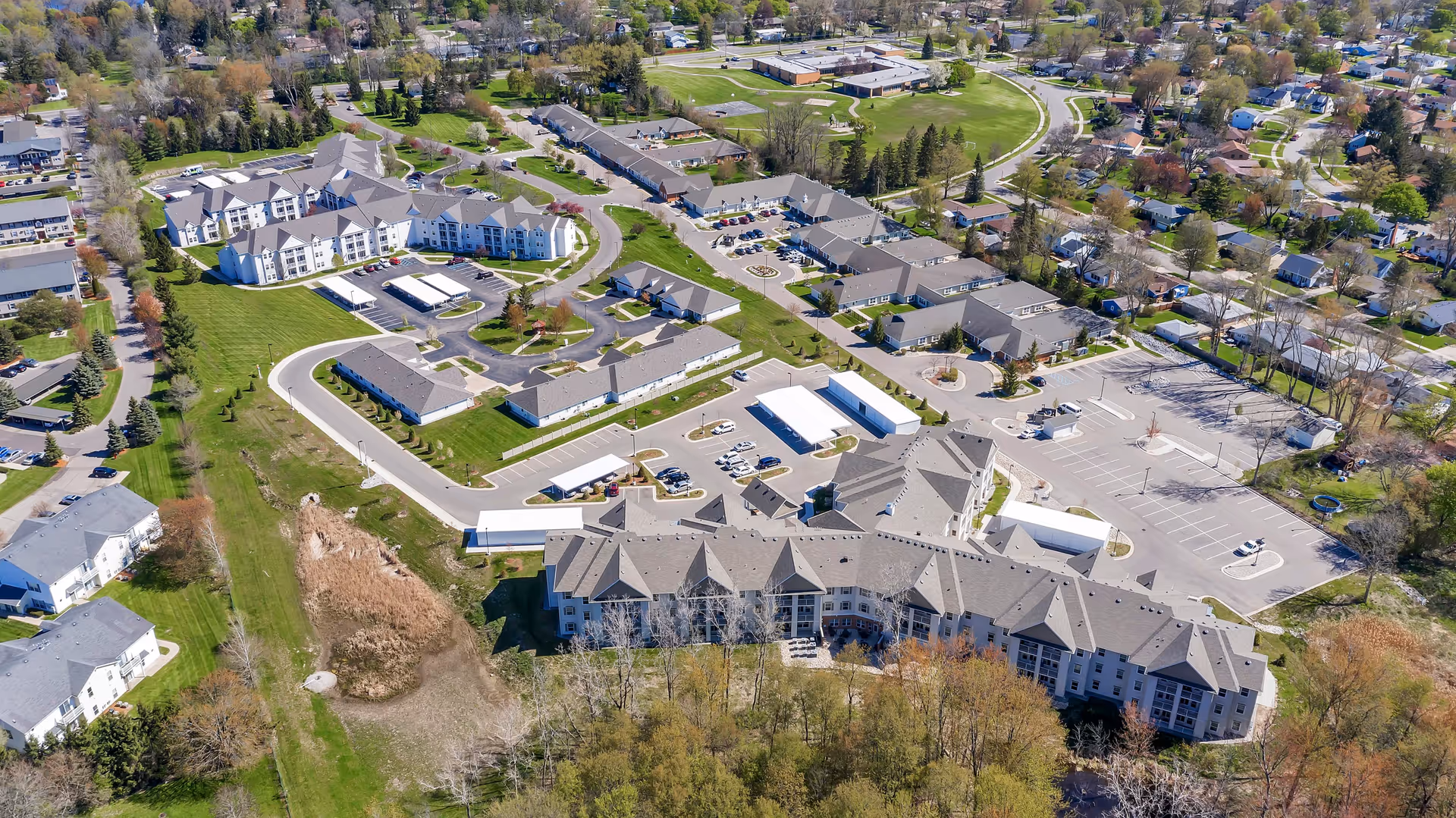 Aerial view of Grandhaven Manor II senior living facility showing multiple buildings with gray roofs, parking lots, driveways, and surrounding green lawns and trees in a suburban neighborhood.