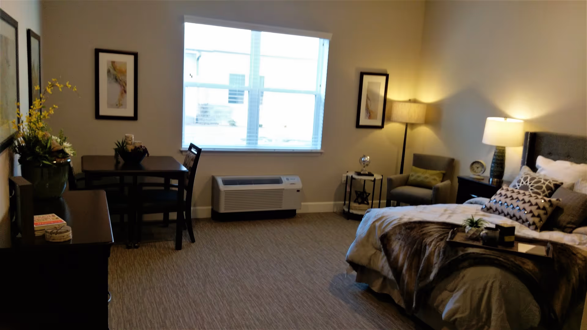 A cozy bedroom in an assisted living facility with a bed adorned with multiple pillows and a throw blanket. There is a nightstand with a lamp and a clock, a comfortable armchair with a floor lamp beside it, and a small table with two chairs near a window. The room is decorated with framed artwork and a vase of flowers on a dresser.