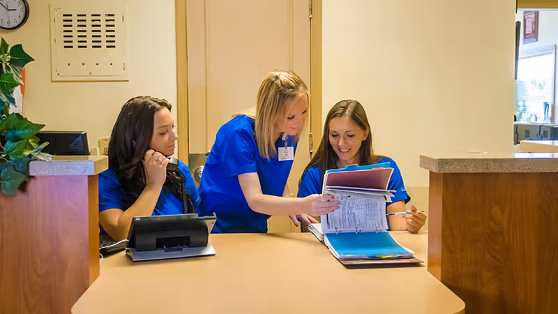 Three women in blue scrubs are at a reception desk. One woman is sitting and looking at a tablet, while the other two are standing and reviewing documents in a binder together. The setting appears to be a healthcare facility reception or nursing station.