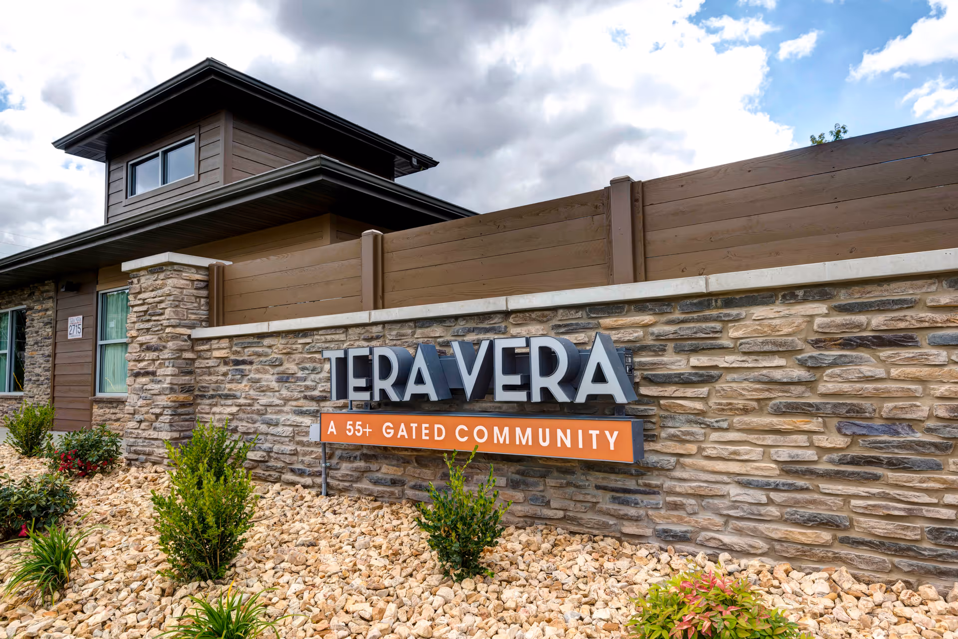 Stone and wood exterior wall with a sign that reads 'TERA VERA A 55+ GATED COMMUNITY' surrounded by small bushes and rocks under a partly cloudy sky.
