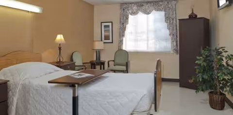 Senior living facility bedroom with a bed, two chairs, window, and tall cabinet.