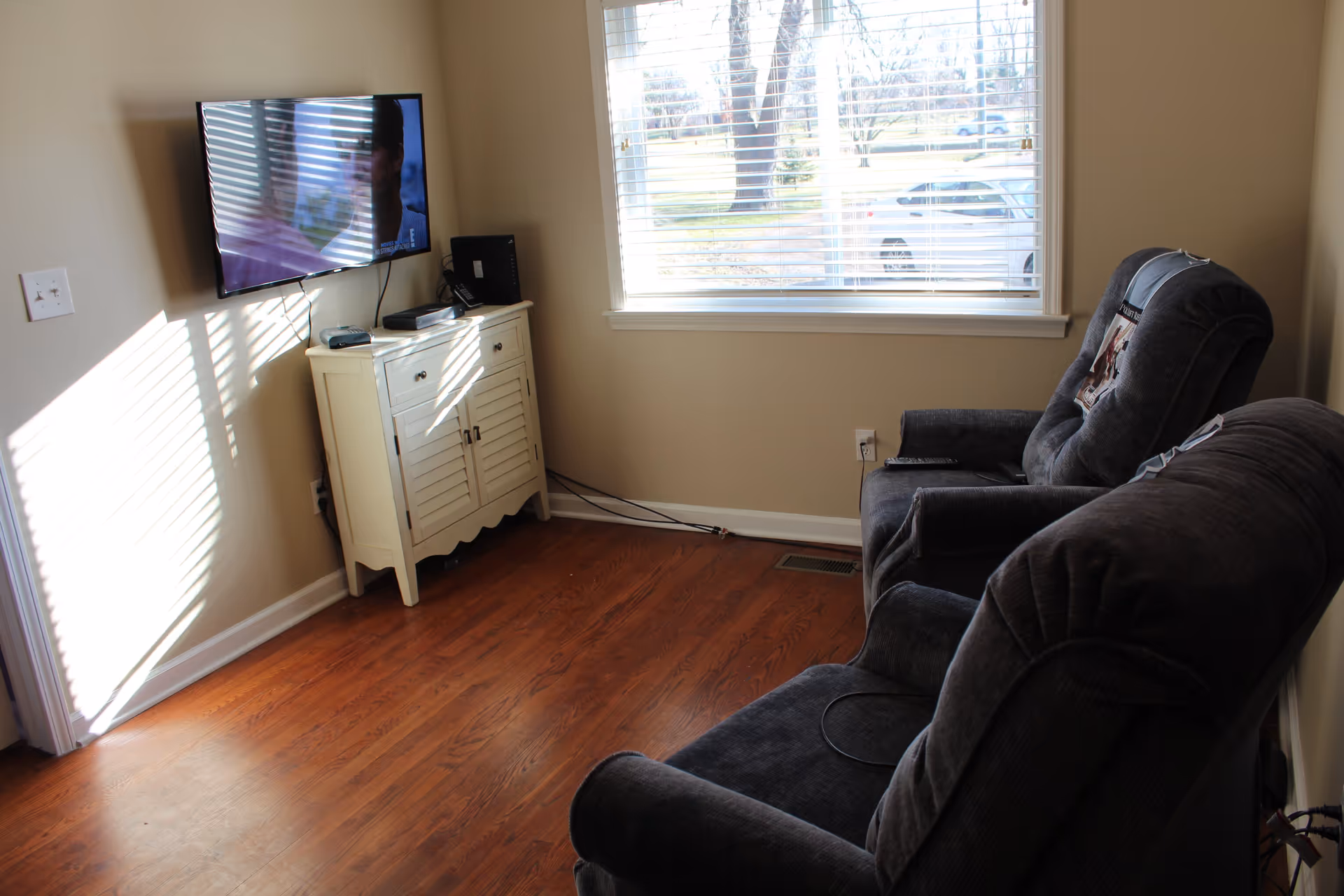 A small living room with two dark gray upholstered recliner chairs facing a wall-mounted flat screen TV. Below the TV is a white cabinet with electronic devices on top. A large window with white blinds lets in natural light, showing a view of trees and parked cars outside. The floor is wooden and the walls are painted beige.