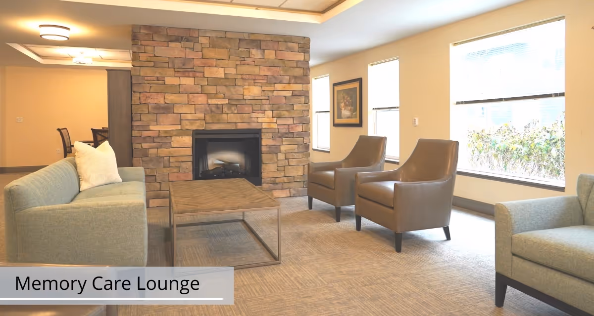 Memory care lounge with sofas, two leather armchairs, a coffee table and a stone fireplace near large windows.