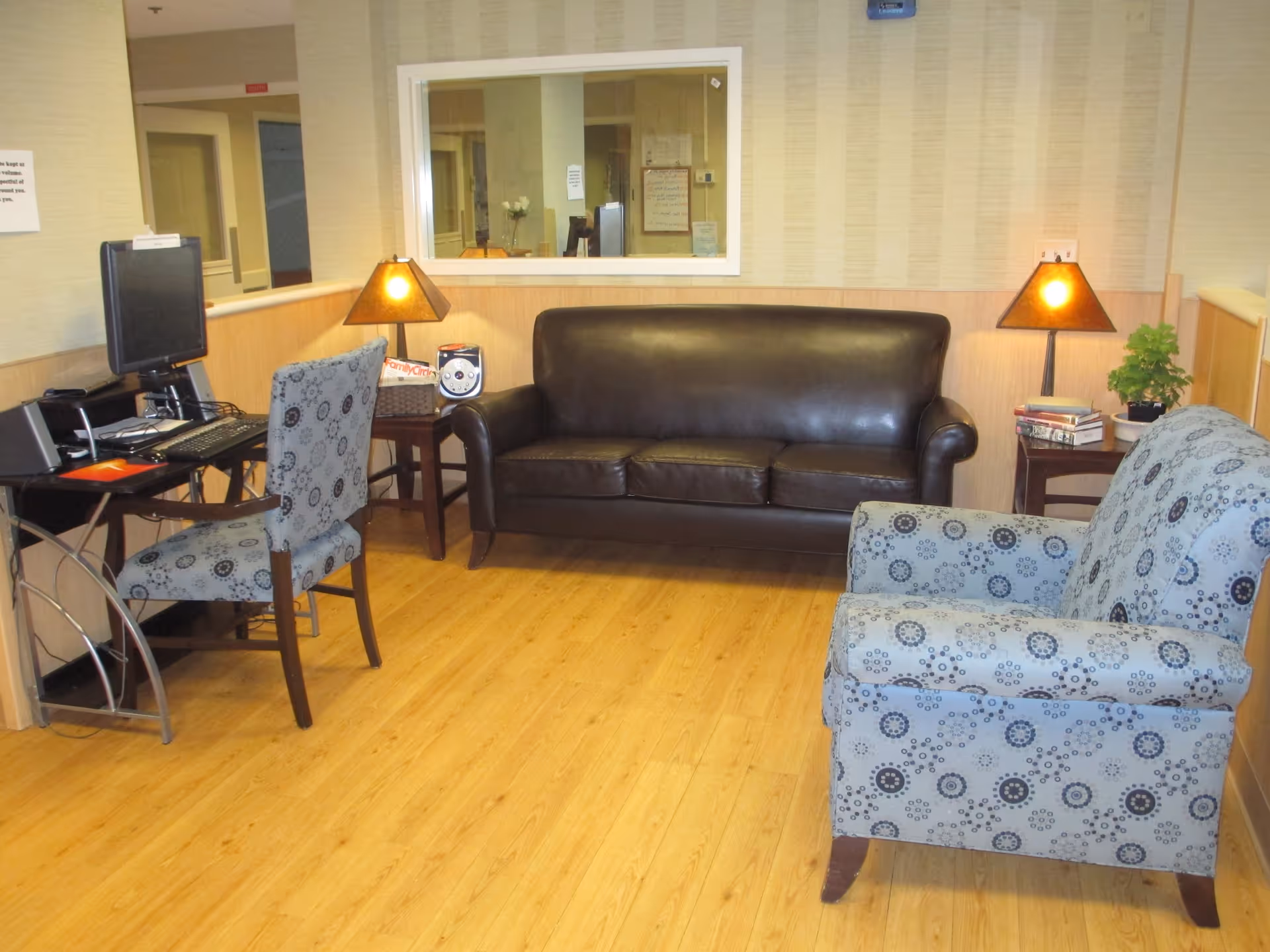 A cozy seating area in a senior living facility with a dark brown leather sofa, a patterned armchair, two side tables each with a lamp, a small potted plant, and a computer workstation with a chair. The room has light wood flooring and beige walls with a large window looking into another room.