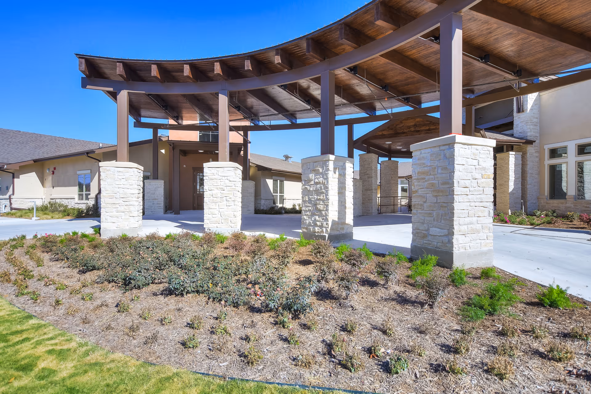 Exterior view of The Brooks of Cibolo Senior Living facility entrance featuring a covered driveway with stone pillars and wooden roof beams, surrounded by landscaped plants and clear blue sky.