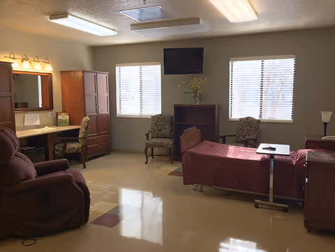 A well-lit room with a hospital-style bed covered in a maroon quilt, two floral upholstered chairs, a recliner, a wooden cabinet, a desk with a mirror and chair, a small bookshelf with a vase of yellow flowers, and a wall-mounted TV between two windows with blinds.