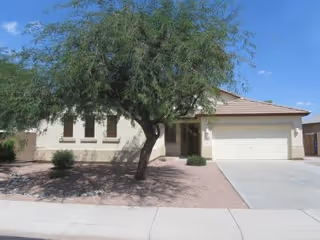 Single-story house with a beige exterior, a large tree in the front yard, gravel landscaping, and a two-car garage under a clear blue sky.