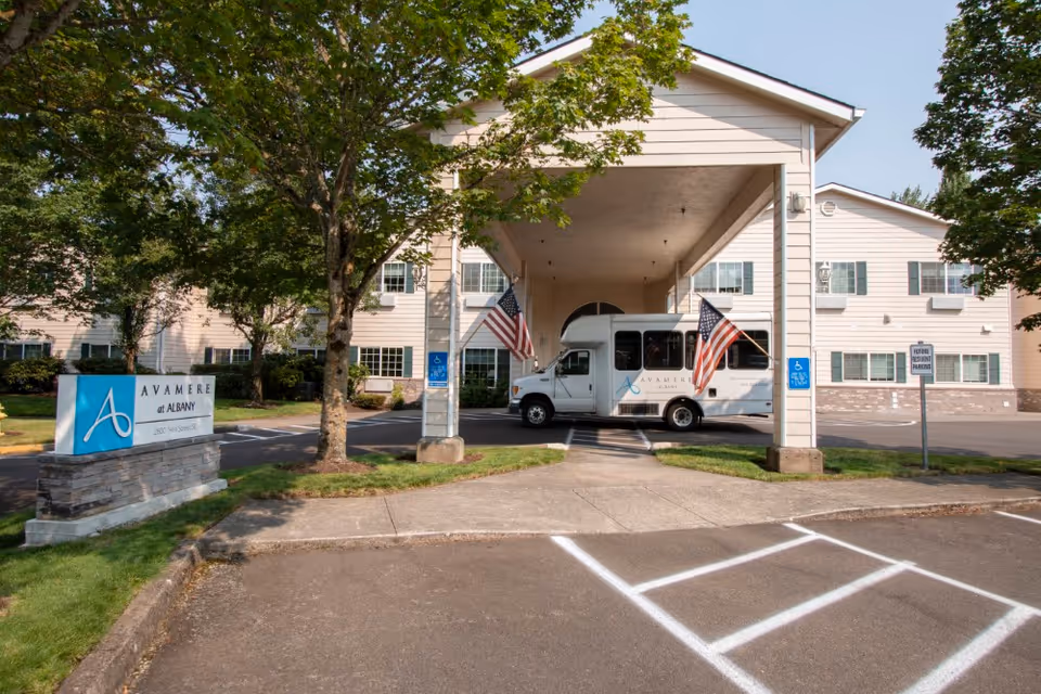 Exterior view of Avamere at Albany senior living facility showing the entrance with a covered drop-off area. A white shuttle van with the Avamere logo is parked under the covered area, flanked by two American flags. There are trees and a sign for Avamere at Albany near the driveway, along with designated handicap parking spaces.