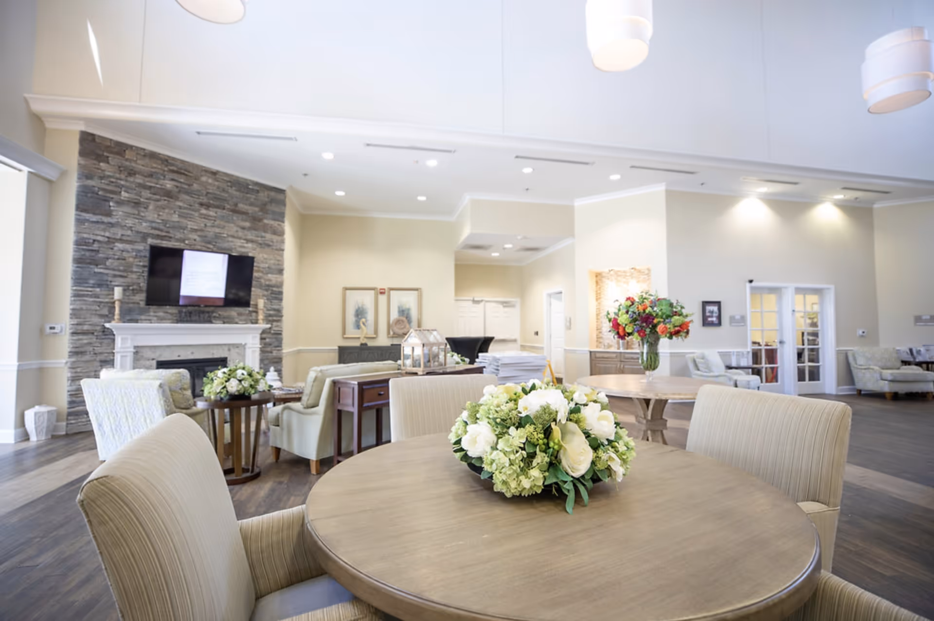 A bright and spacious living room area in a senior living facility featuring a round wooden table with a floral centerpiece in the foreground, comfortable upholstered chairs, a stone fireplace with a mounted flat-screen TV, additional seating areas with armchairs, and decorative flowers on another table in the background.