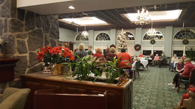A cozy common area in a retirement community decorated for the holidays with a Christmas tree and wreaths. Several elderly people are seated and standing, engaging in conversation. The room features large arched windows, chandeliers, a stone fireplace, and green carpeted flooring.