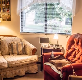 Cozy living room with a patterned sofa and a red recliner beside a window, and a side table holding a lamp and telephone.