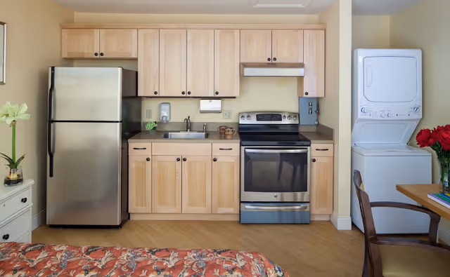 A compact kitchen area with light wood cabinets, a stainless steel refrigerator, an electric stove with oven, and a sink. To the right, there is a stacked washer and dryer unit next to a wooden table with a chair and a vase of red flowers. The floor is light wood, and part of a bed with a floral bedspread is visible in the foreground.