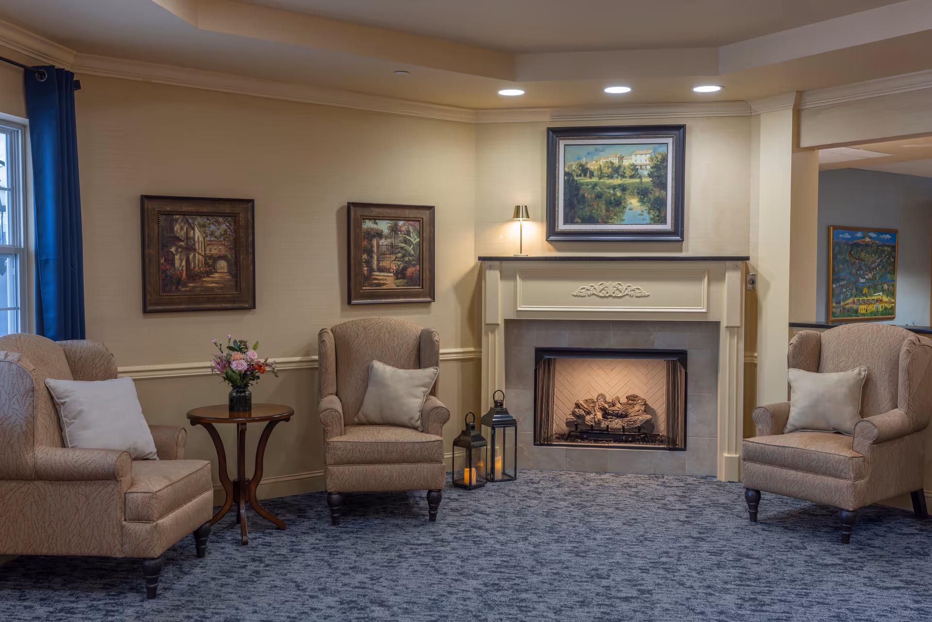 A cozy living room area with three beige upholstered armchairs, each with a light-colored pillow. There is a small wooden side table with a vase of flowers between two of the chairs. The room features a fireplace with a decorative mantel and a framed painting above it. Two framed paintings hang on the wall to the left of the fireplace. The floor is carpeted in a patterned blue-gray color, and there are two lantern-style candle holders on the floor next to the fireplace. The room is softly lit with recessed ceiling lights and a small lamp on the mantel.