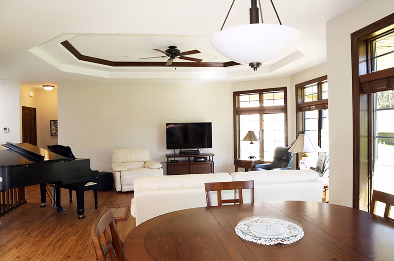 Bright living room with large windows, white leather sofas, a wooden dining table with chairs, a black grand piano, a ceiling fan, and a flat-screen TV on a wooden stand.