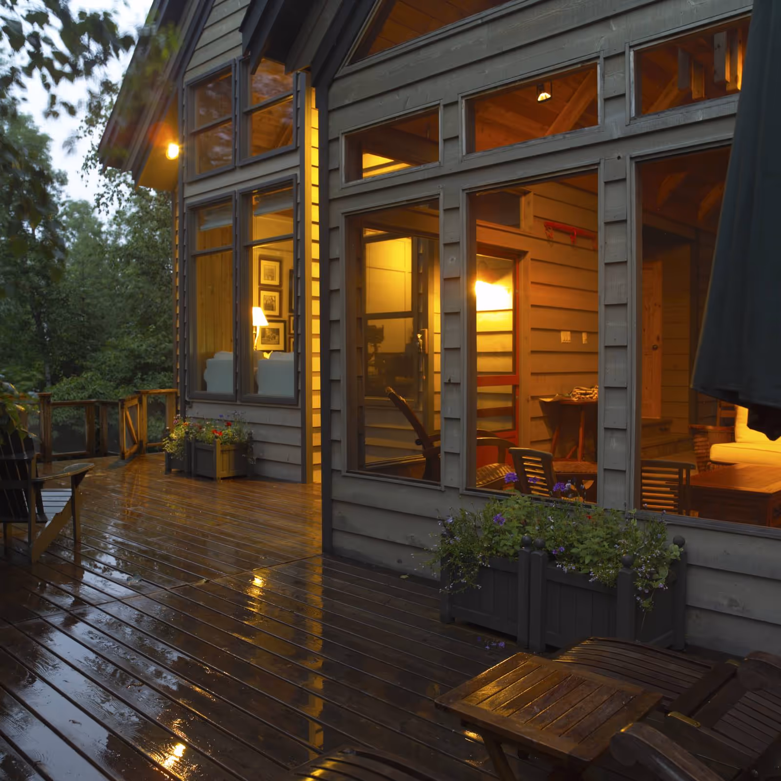 Rain-wet wooden deck outside a house with large windows revealing warmly lit interior rooms and potted planters.
