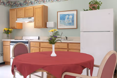 A small kitchen area with wooden cabinets, a white refrigerator, a stove with a range hood, and a sink. In the foreground, there is a round table covered with a red tablecloth and a white vase with flowers. The walls are light green with a floral wallpaper border near the ceiling, and a framed picture hangs above the sink.