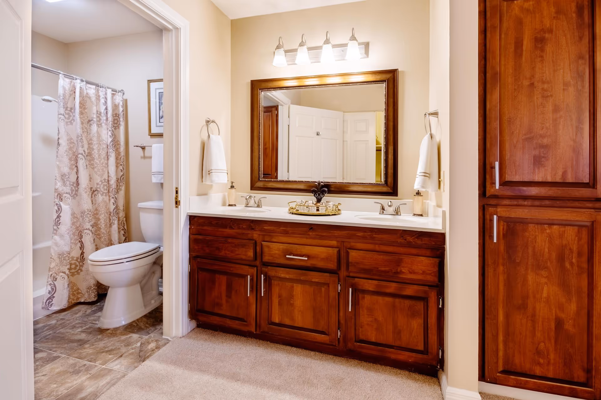 A bathroom with a wooden double-sink vanity and large mirror, and a toilet and shower with a patterned curtain.