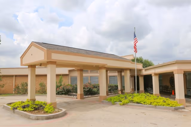 Entrance area of a single-story building with a covered drop-off zone supported by columns, landscaped with green plants and bushes, and an American flag on a flagpole under a cloudy sky.