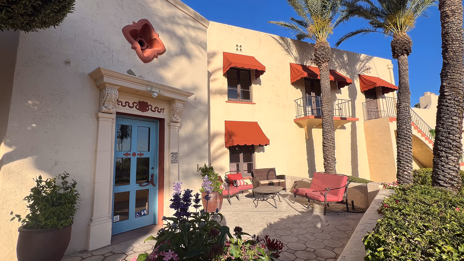 Exterior view of a senior living facility with a light yellow stucco wall, blue door with decorative molding, red window awnings, and outdoor seating with red cushions. There are palm trees and potted plants in the foreground under a clear blue sky.