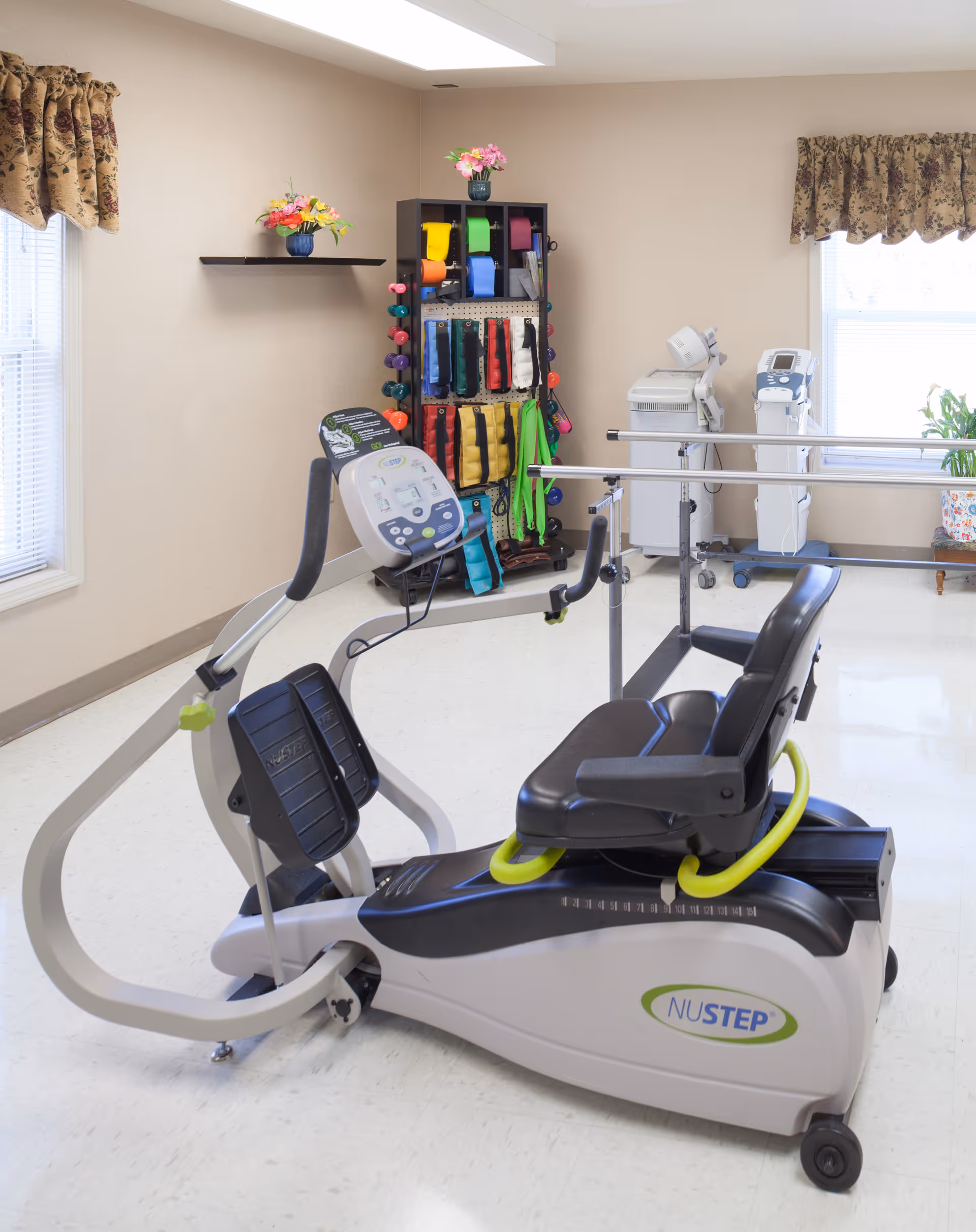 Recumbent NuStep exercise machine and parallel bars in a bright therapy/exercise room with shelves of colorful resistance bands and windows with floral valances.