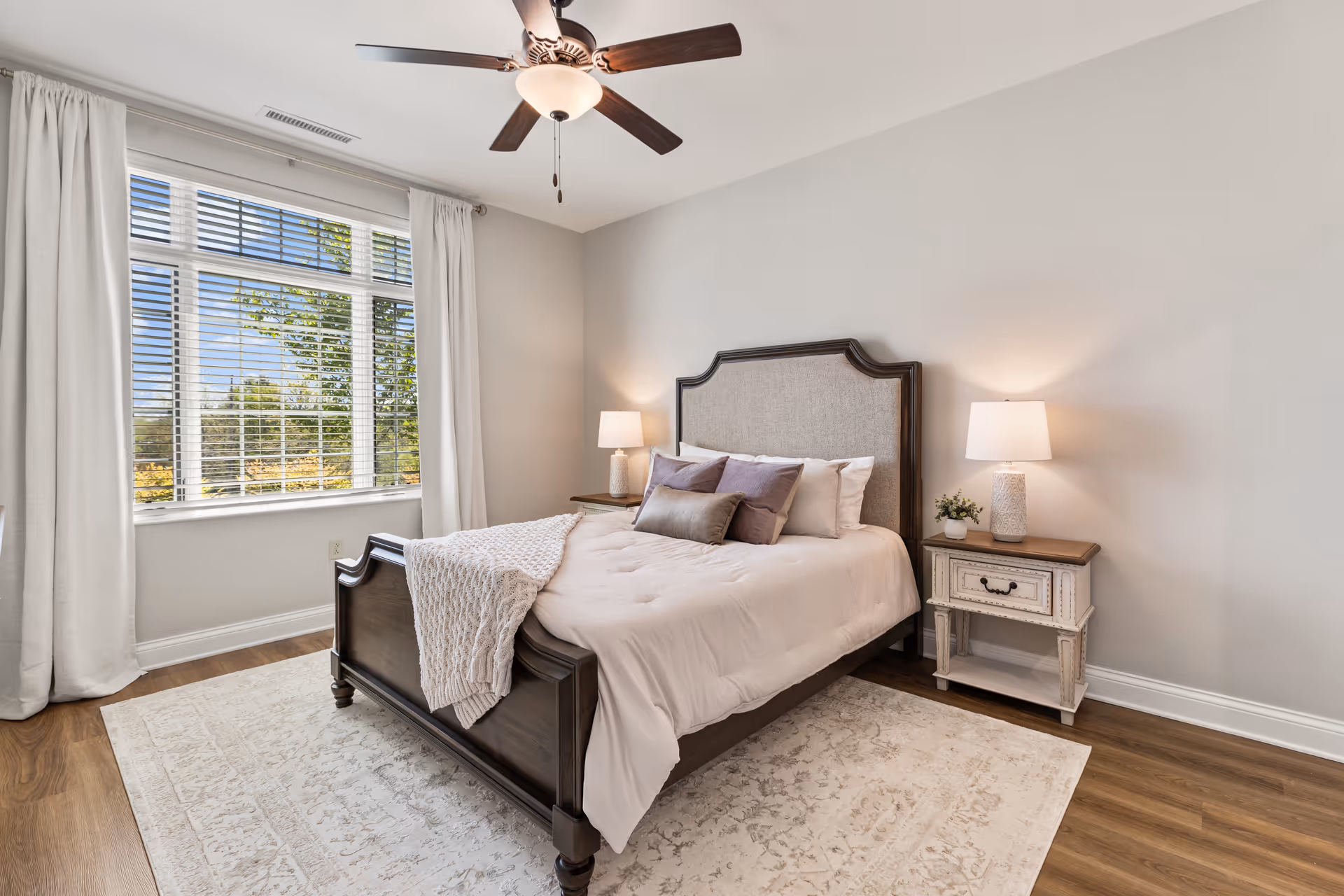 A bright and cozy bedroom with a large window covered by white curtains and blinds, a wooden bed with a cushioned headboard, white bedding with several pillows, a knitted throw blanket, two bedside tables each with a lamp, a small plant on one table, a ceiling fan with a light fixture, and a light-colored rug on wooden flooring.