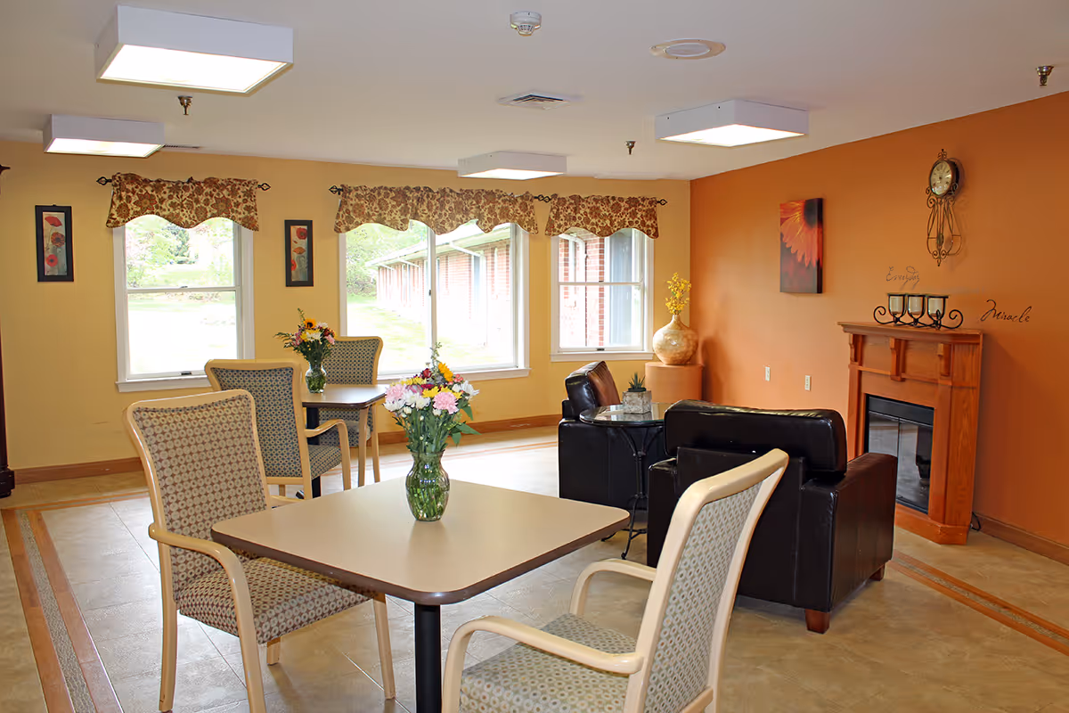 A cozy common area in a senior living facility with a square table and four patterned chairs, a vase with flowers on the table, two black leather armchairs around a small glass table, a wooden fireplace with candles and a clock above it, orange and yellow walls, and windows with floral valances letting in natural light.
