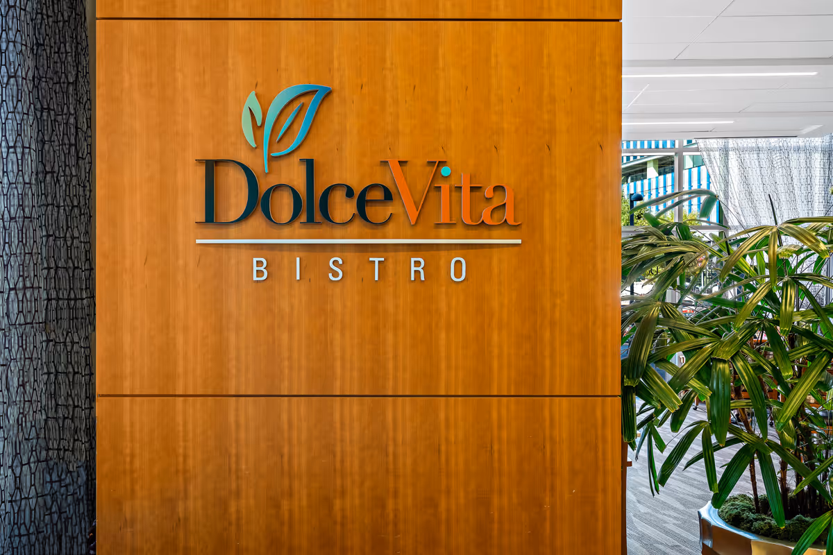 A wooden wall with the sign 'Dolce Vita Bistro' mounted on it, featuring a leaf logo above the text. To the right, there are green plants and part of an interior space with large windows and curtains.