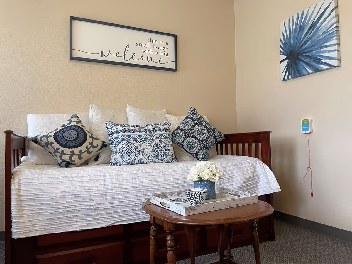 A cozy living room area with a wooden daybed covered in a white textured blanket and decorated with multiple blue and white patterned pillows. Above the daybed is a framed sign that reads 'this is a small house with a big welcome.' A small wooden coffee table in front of the daybed holds a white tray with a blue vase of white flowers and a patterned cup. On the adjacent wall, there is a blue and white floral artwork and a medical alert device mounted with a red pull cord hanging down.