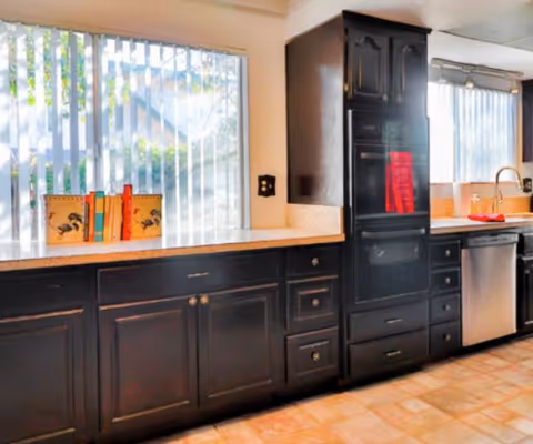 Sunlit kitchen with dark wood cabinets and a tall pantry, light countertops, a sink and stainless dishwasher beneath windows with vertical blinds.