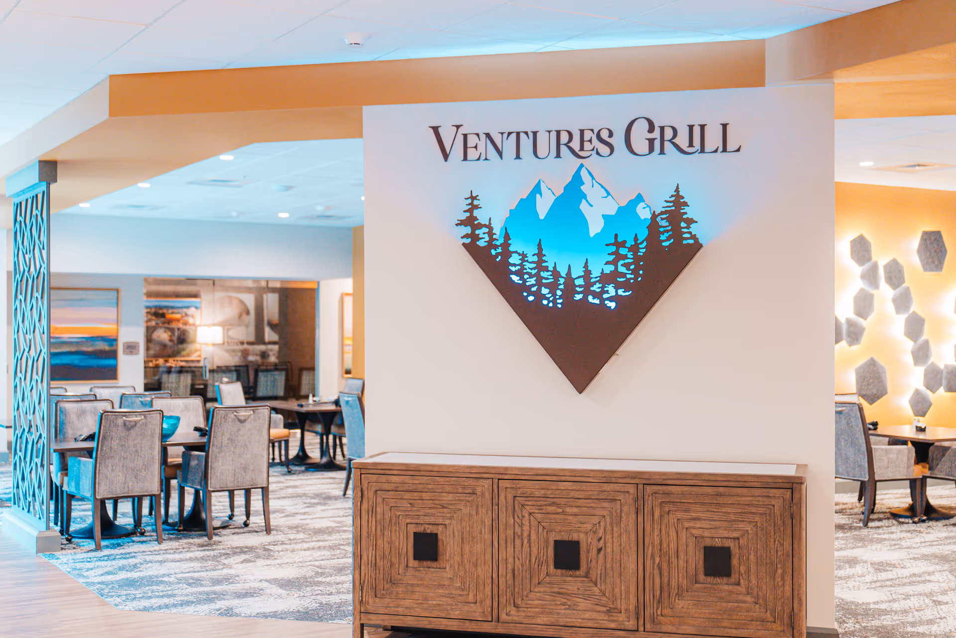 Interior view of a dining area named Ventures Grill with multiple tables and chairs arranged for seating. The space features a decorative sign with a mountain and tree design on the wall, soft lighting, and modern decor.