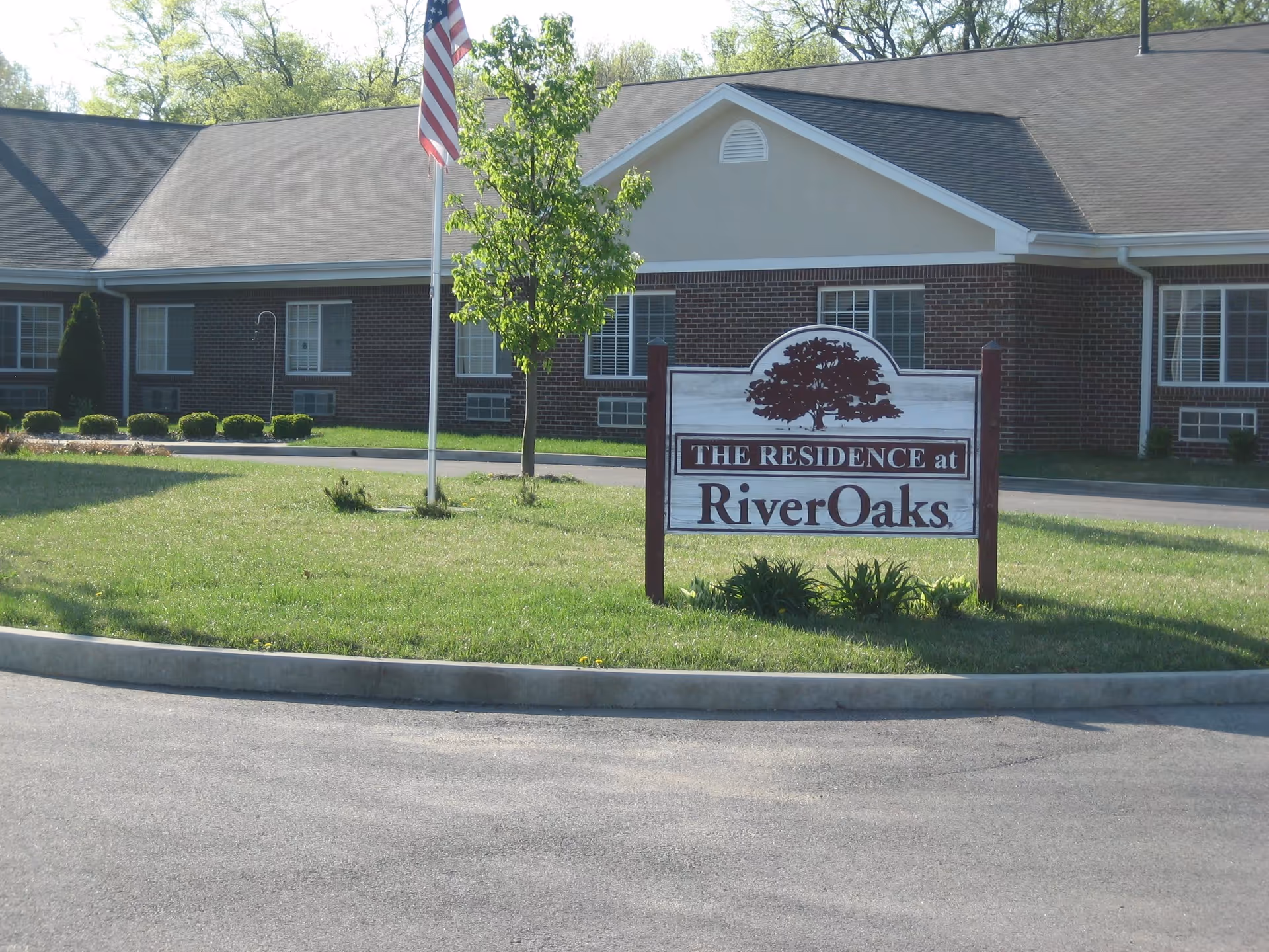 Exterior view of a single-story brick building with a sign in front that reads 'THE RESIDENCE at RiverOaks'. There is a small tree and an American flag on a flagpole near the sign, with a paved driveway and green grass surrounding the area.