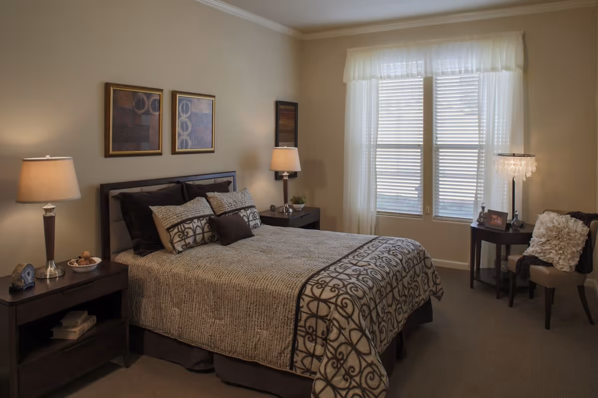 A cozy bedroom with a neatly made bed featuring patterned bedding and multiple pillows. There are two bedside tables with lamps on either side of the bed. The room has a window with sheer white curtains allowing natural light to enter. A small round table with a lamp and a chair with a decorative pillow are placed near the window. The walls are decorated with framed artwork.