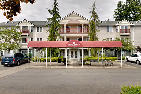 Front exterior view of a three-story senior living facility with beige siding and red balconies. There is a red canopy over the entrance with the text 'Merrill Gardens' and a logo. Several cars are parked in front, and there are trees and shrubs around the building.