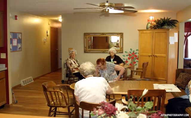 A group of elderly women sitting and interacting around a wooden dining table in a cozy room with wooden floors, a ceiling fan, a large mirror on the wall, and a wooden cabinet with a lamp and plants on top.