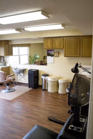Interior of a hair salon area with a pink salon chair, a wall-mounted sink, wooden cabinets, a calendar on the wall, two laundry baskets, and a hair dryer chair. The floor is wooden, and there is a window letting in natural light.