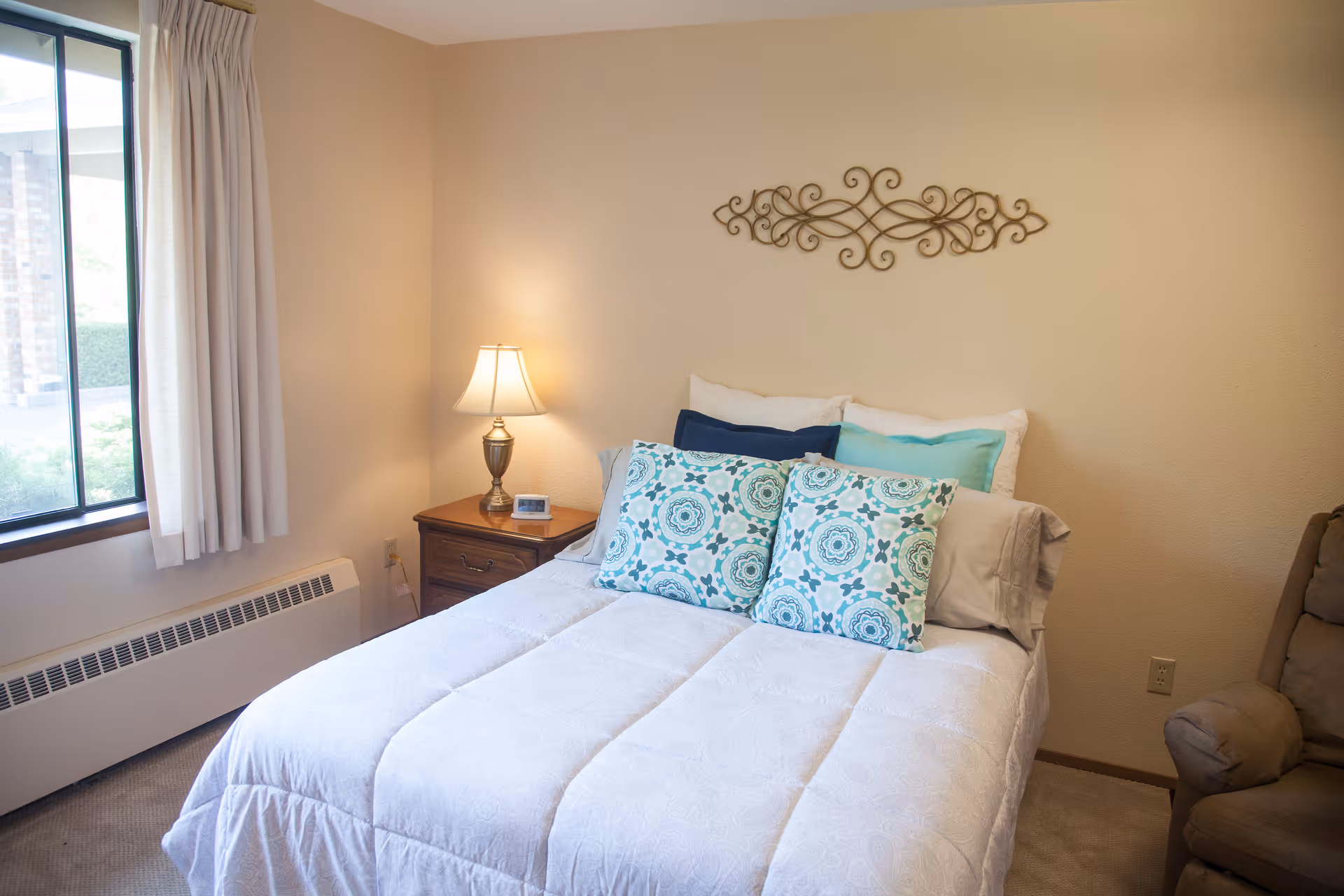 A cozy bedroom with a neatly made bed featuring white bedding and decorative blue and turquoise pillows. There is a wooden nightstand with a lamp and a small clock next to the bed. A window with beige curtains lets in natural light, and a decorative metal wall art piece hangs above the bed. A cushioned armchair is partially visible on the right side.