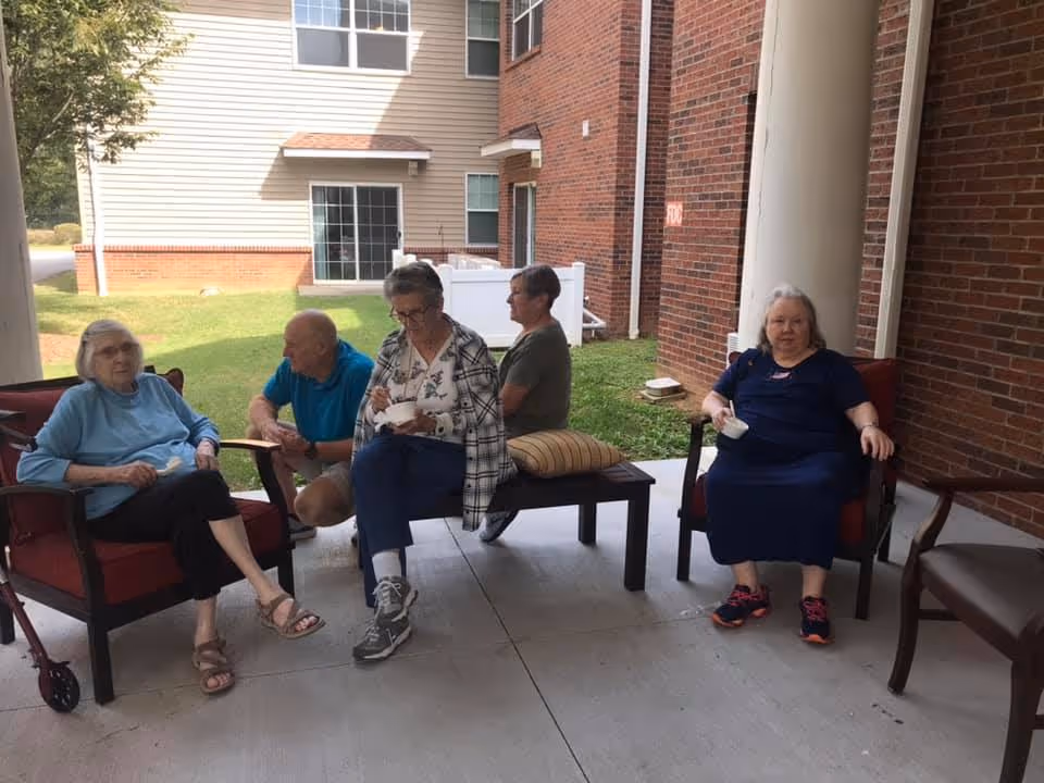 Five elderly people sitting and socializing on a covered patio area outside a brick and siding building. Two women sit in armchairs, one man crouches near a woman eating from a bowl, and another woman sits on a bench with a cushion behind her. The outdoor area has concrete flooring and overlooks a grassy yard.