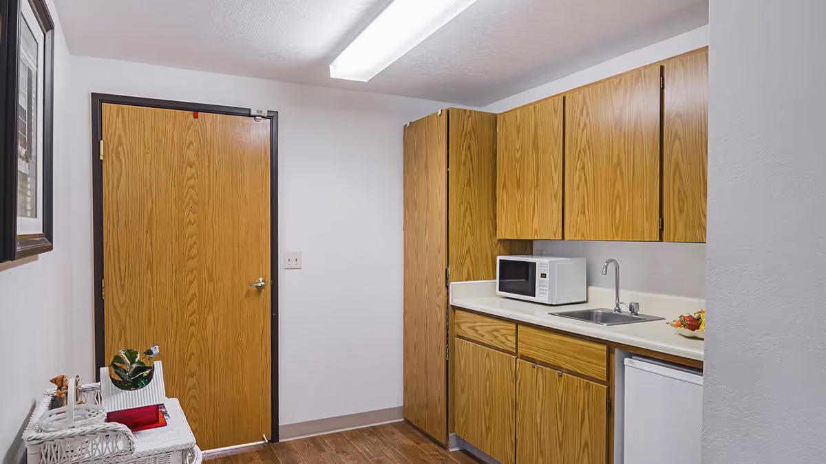 Small kitchenette with wood cabinets, a countertop sink, microwave and mini-refrigerator next to a closed wooden door.