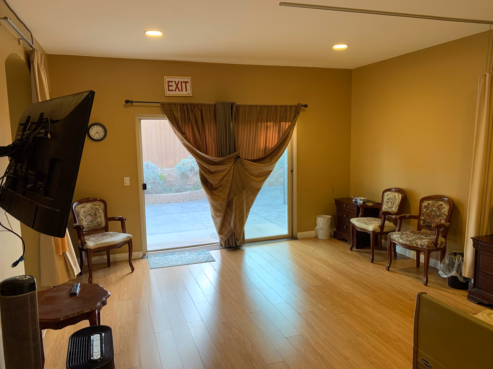 Empty common living room with wood floors, patterned chairs, a wall-mounted TV, and a sliding glass door with curtains tied in the center.