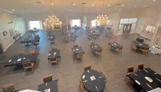Spacious dining room with multiple tables and chairs arranged neatly. The room features large windows allowing natural light, chandeliers hanging from the ceiling, and a piano in the corner. The flooring is wood, and the walls are painted in light colors, creating a bright and welcoming atmosphere.