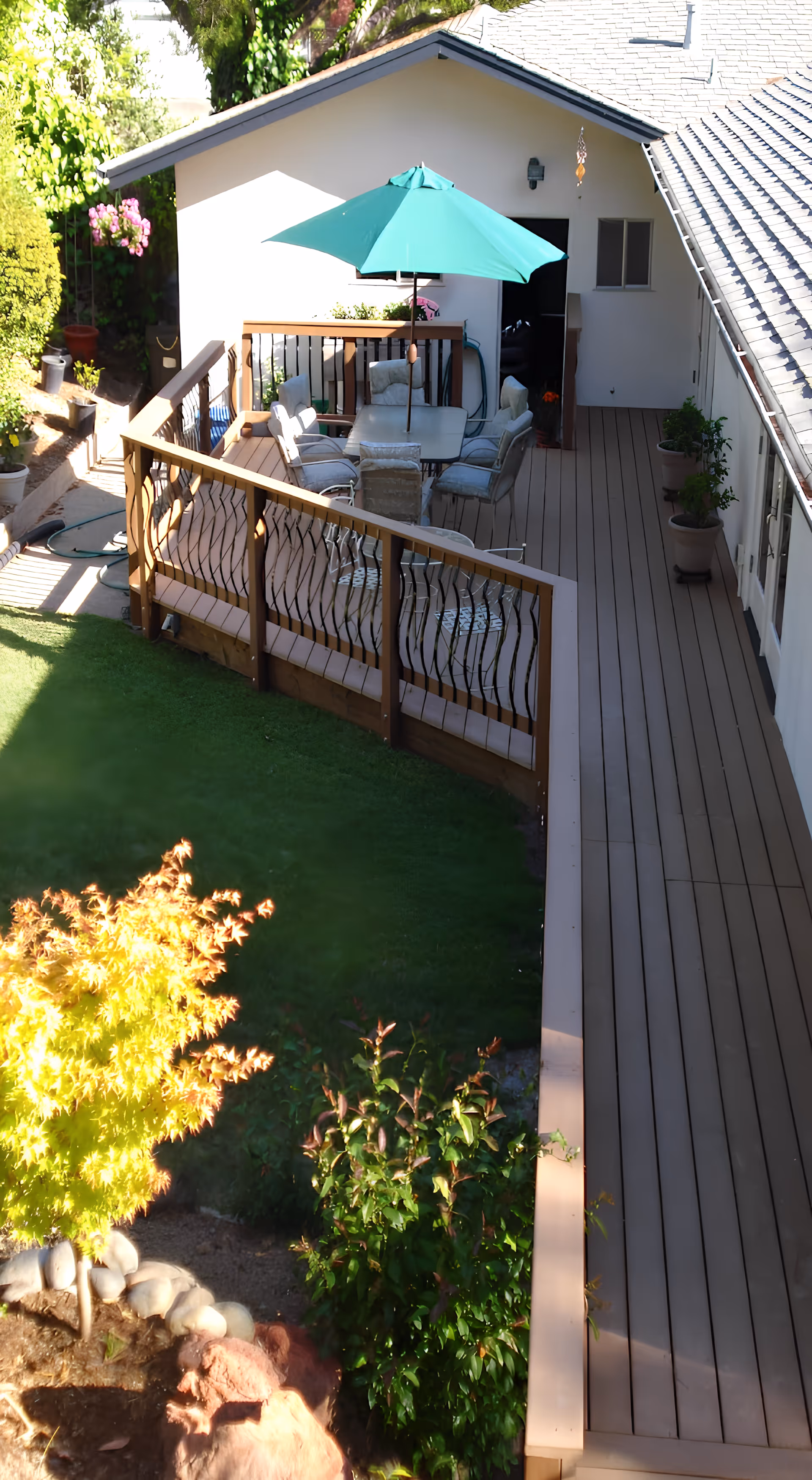 Outdoor patio area with a wooden deck attached to a white building. The deck has a table with six cushioned chairs and a large green umbrella providing shade. Several potted plants line the deck, and a garden with green grass, bushes, and a small tree with yellow leaves is visible adjacent to the deck.